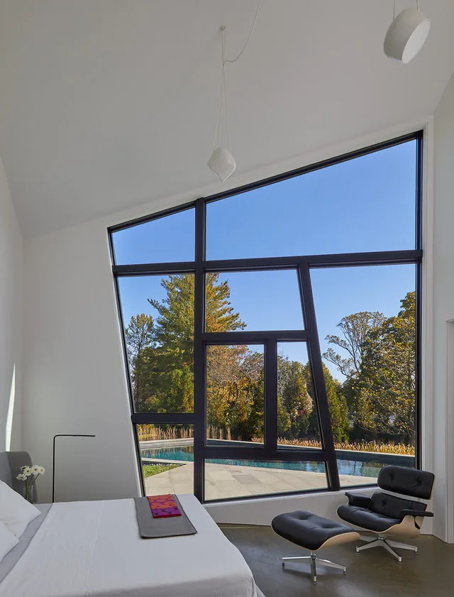 Bedroom with a large angular window looking out to a pool and autumn trees during daylight.