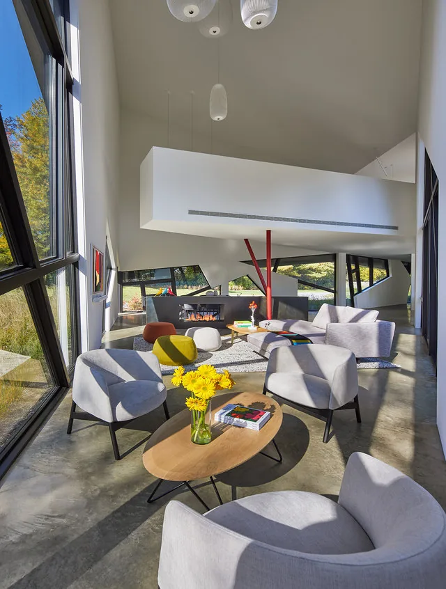 PTR HOUSE living room features angular window design and built-in shelving around a central fireplace with mesh railing and bright seating.