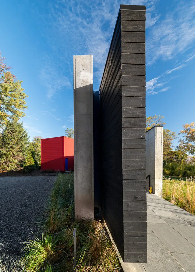 PTR HOUSE by Robert Gurney Architects features contrasting dark wood siding and concrete walls framing an outdoor path with natural planting.