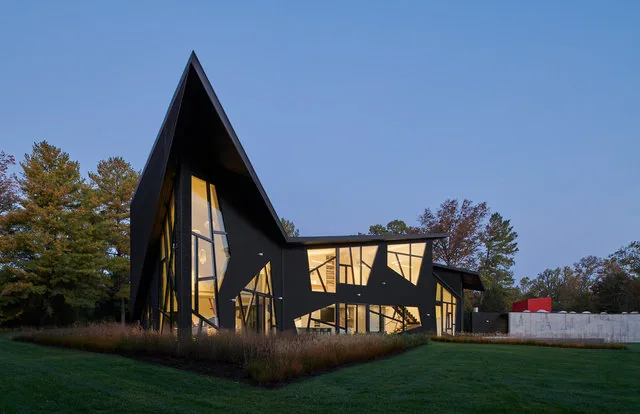 PTR HOUSE features an angular roof and asymmetrical windows with a sleek dark exterior and illuminated interior at dusk.