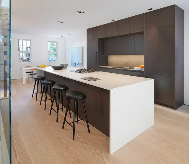 A contemporary kitchen design features a large white island with black stools and dark wood cabinetry along the wall.