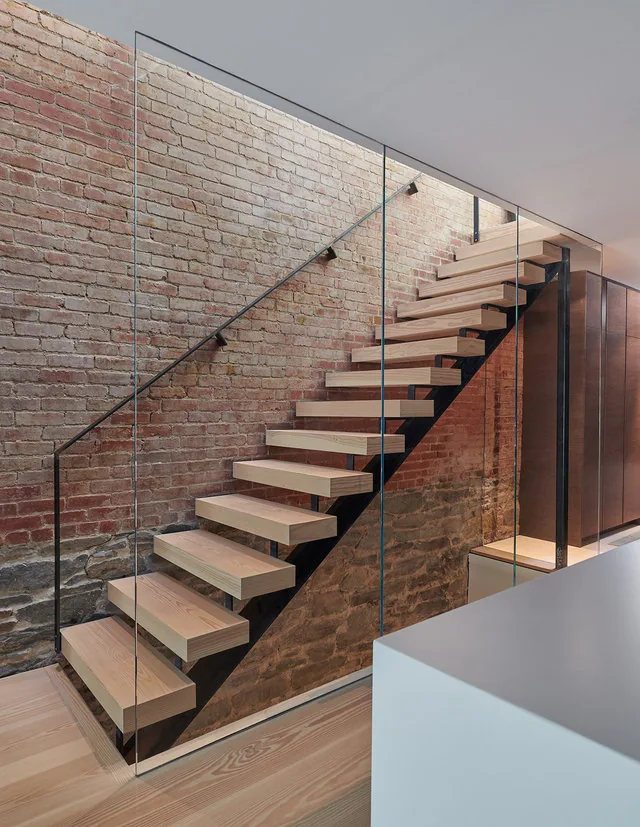 Floating wood stairs with glass railing and exposed brick wall in an interior space.