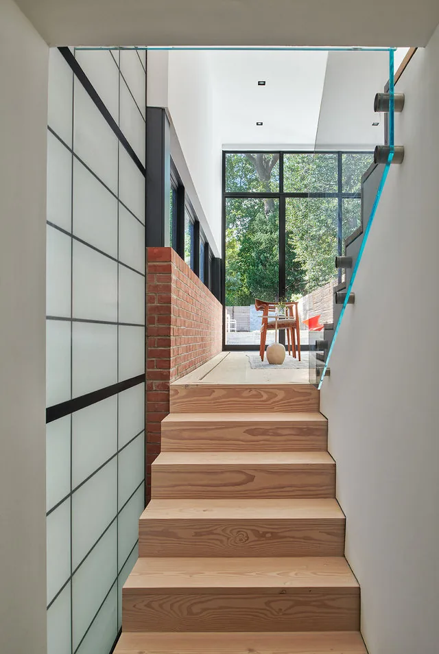 Modern wood stair with glass railing leads to a living space with large black-framed windows.