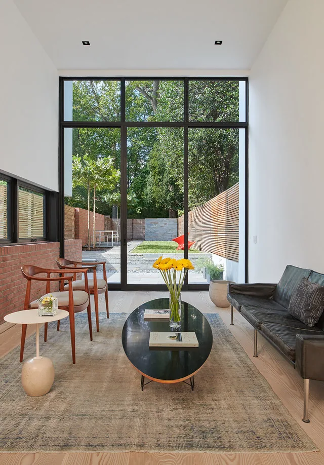 Living room features floor-to-ceiling black window frames overlooking a fenced backyard with a red outdoor sculpture.