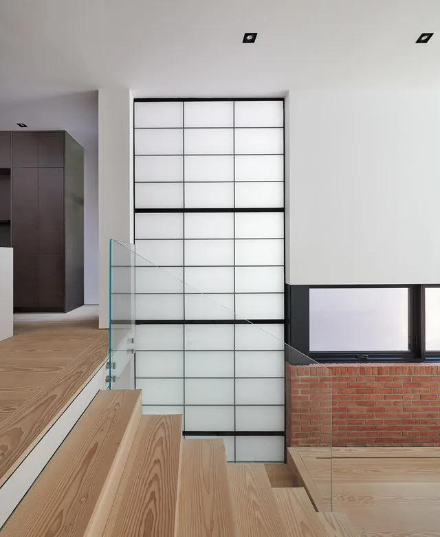 Wood stairs with glass railing beside a large shoji-style window and exposed brick wall.