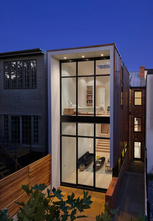 A modern renovation with large black window frames reveals a double-height living room and upstairs bedroom, with wood stairs and minimal décor.