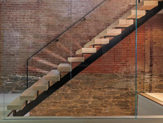 Floating wood staircase with a glass railing is set against an exposed brick and stone wall.