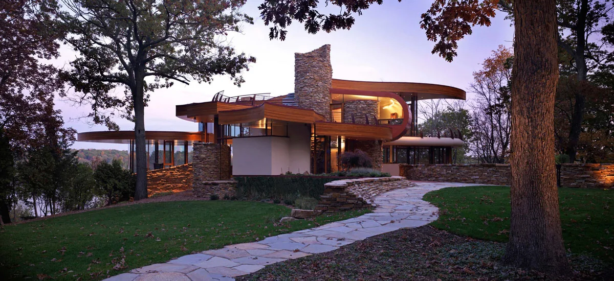 Chenequa Residence features curved wooden rooflines and stone walls with expansive glazing, set in a wooded landscape.