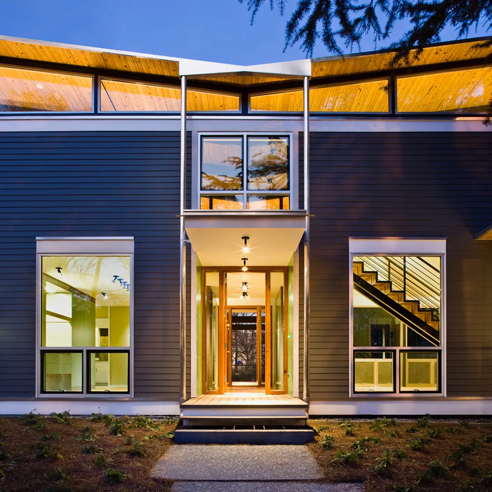 Illuminated modern house front with gray horizontal wood siding and large windows showing interior stair and kitchen area.