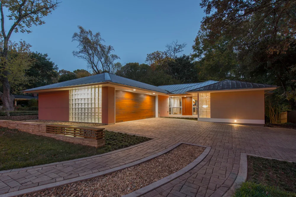 Midcentury modern house exterior with glass block, textured garage door, and metal roof, set in a wooded landscape at dusk.