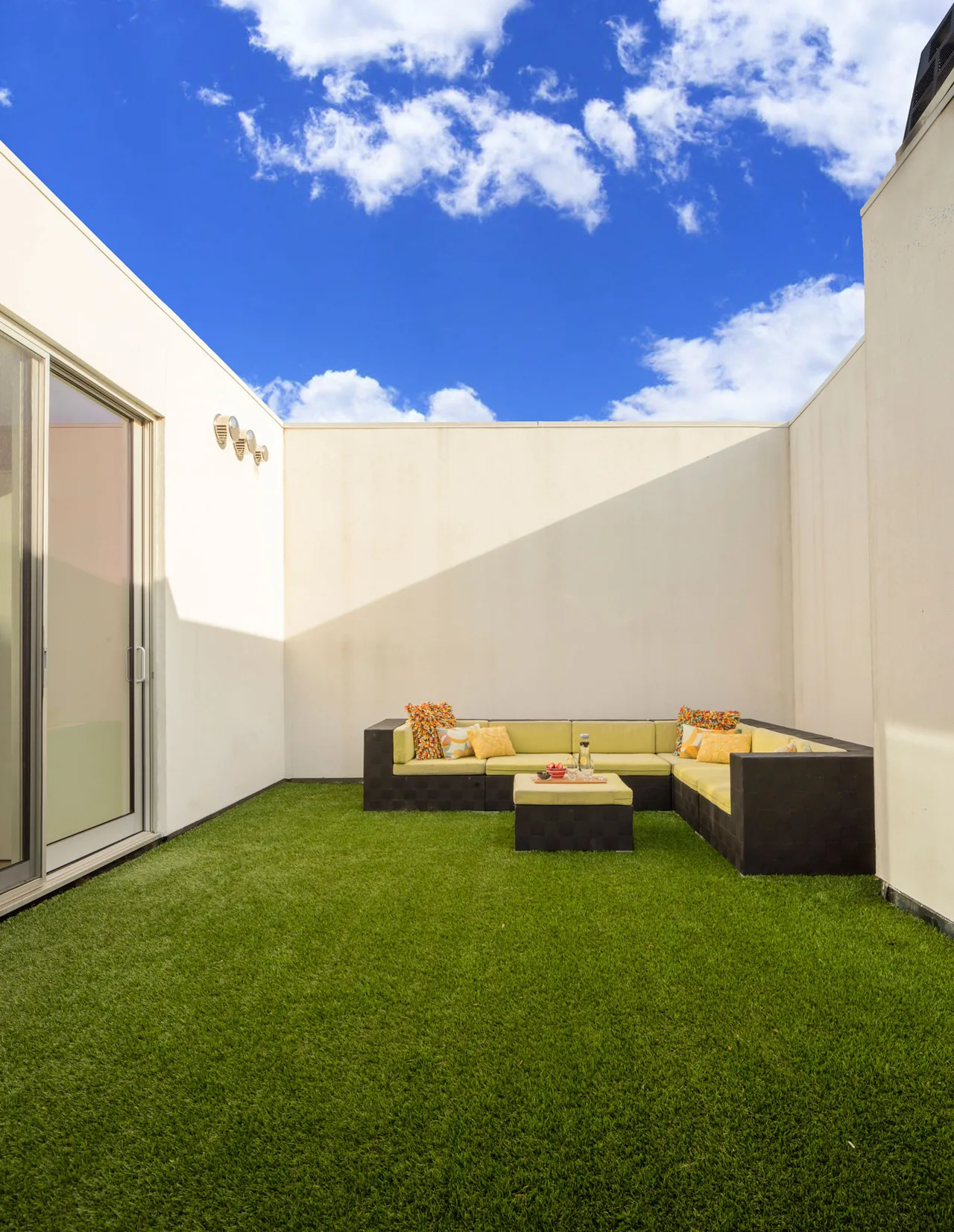 House 432 courtyard with artificial grass and sectional seating under a sky with scattered clouds.