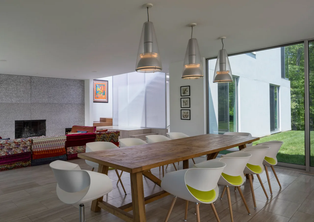 Dining area in House 432 features a wooden table with modern white chairs and pendant lighting, with floor-to-ceiling windows opening to the yard.