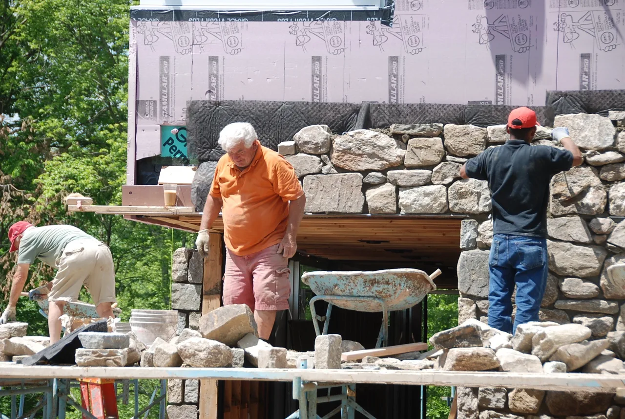 Three workers constructing a rough stone wall as part of an outdoor building under construction.