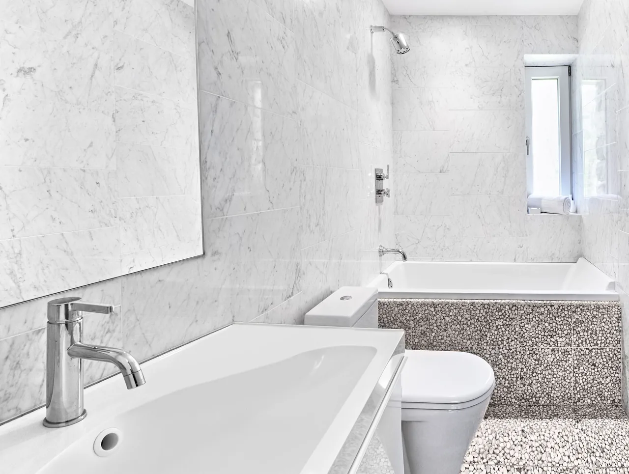 Bathroom interior featuring white marble walls, pebble tile floor and tub surround, and a white tub beneath a wall-mounted showerhead.