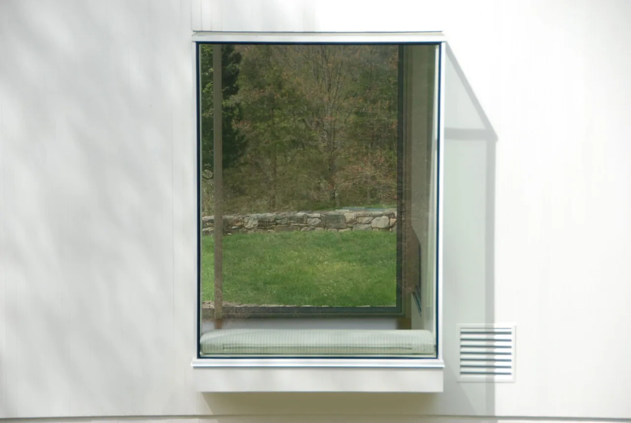 A square window of House 125 offers a view of the garden's stone retaining wall and grass from a built-in bench seat.