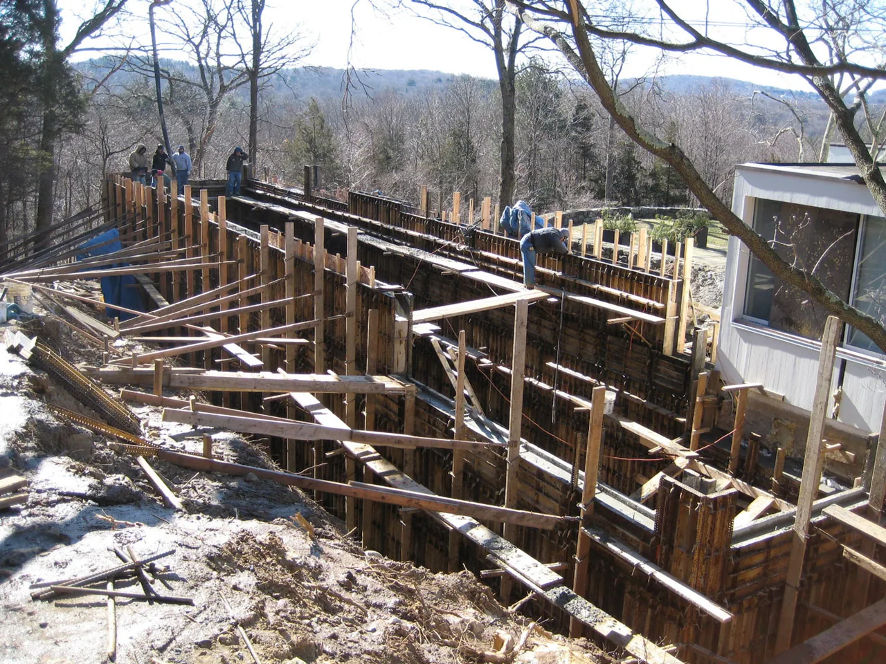 Workers build wood formwork for a retaining wall on a sloped site adjacent to an existing house structure.