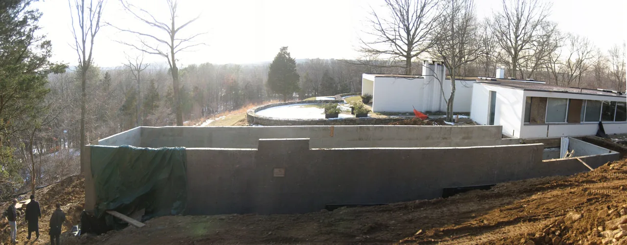 House 125 features a flat-roofed modern design with a prominent concrete retaining wall on a wooded sloped site under construction.