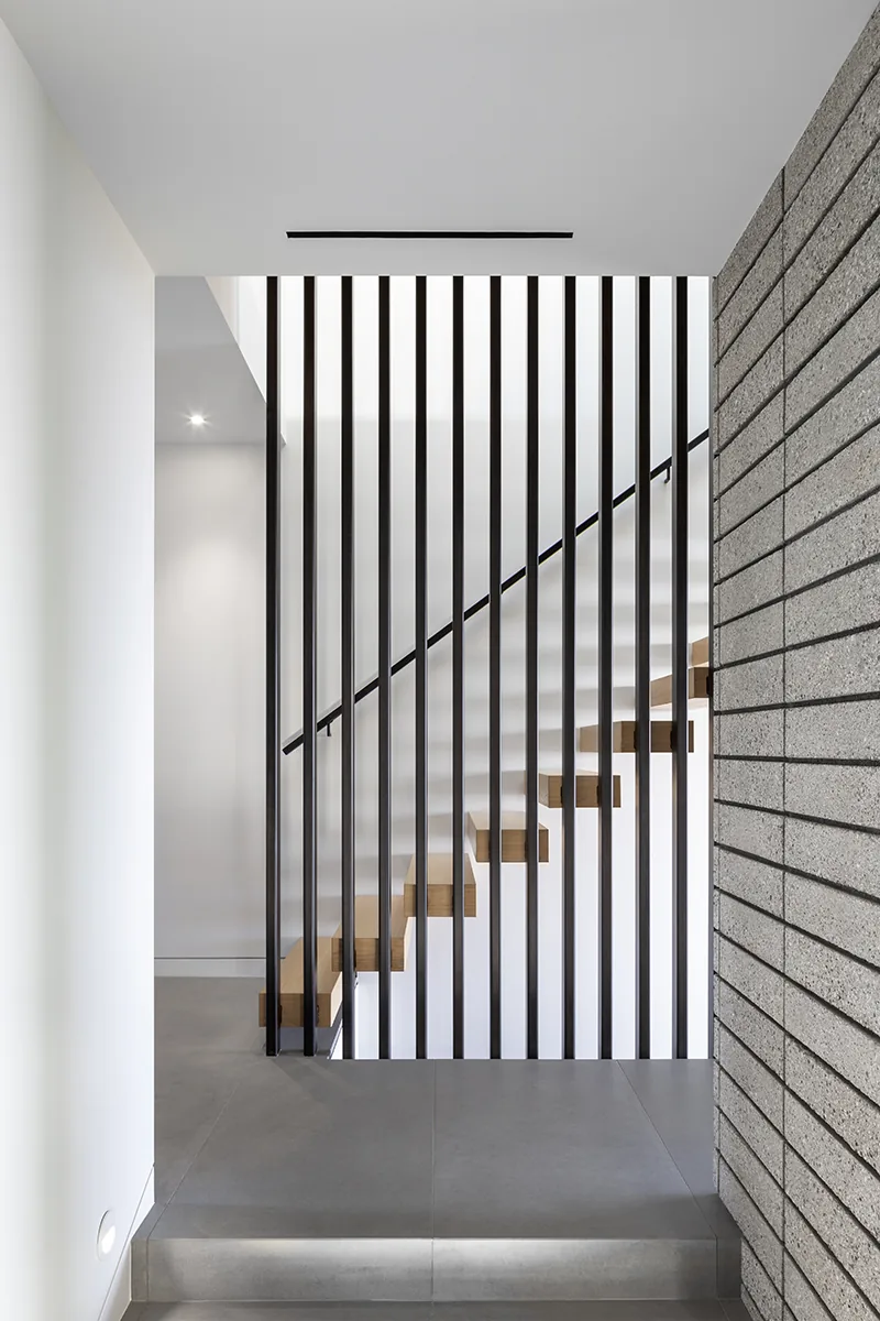 Floating wooden stairs feature vertical black metal balusters, adjacent to a textured gray brick wall and gray tile floor.