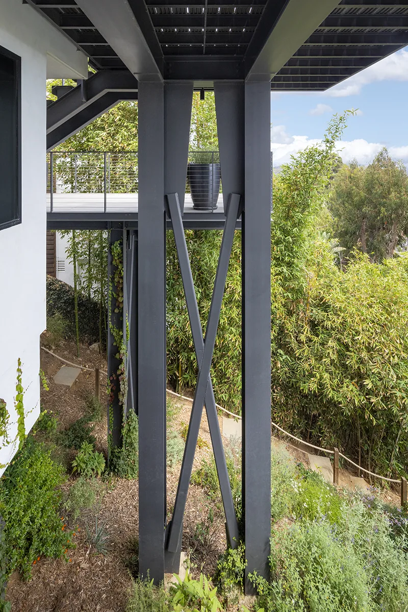 Black metal columns and cross braces support an elevated deck with metal cable railing, overlooking landscaped greenery below.