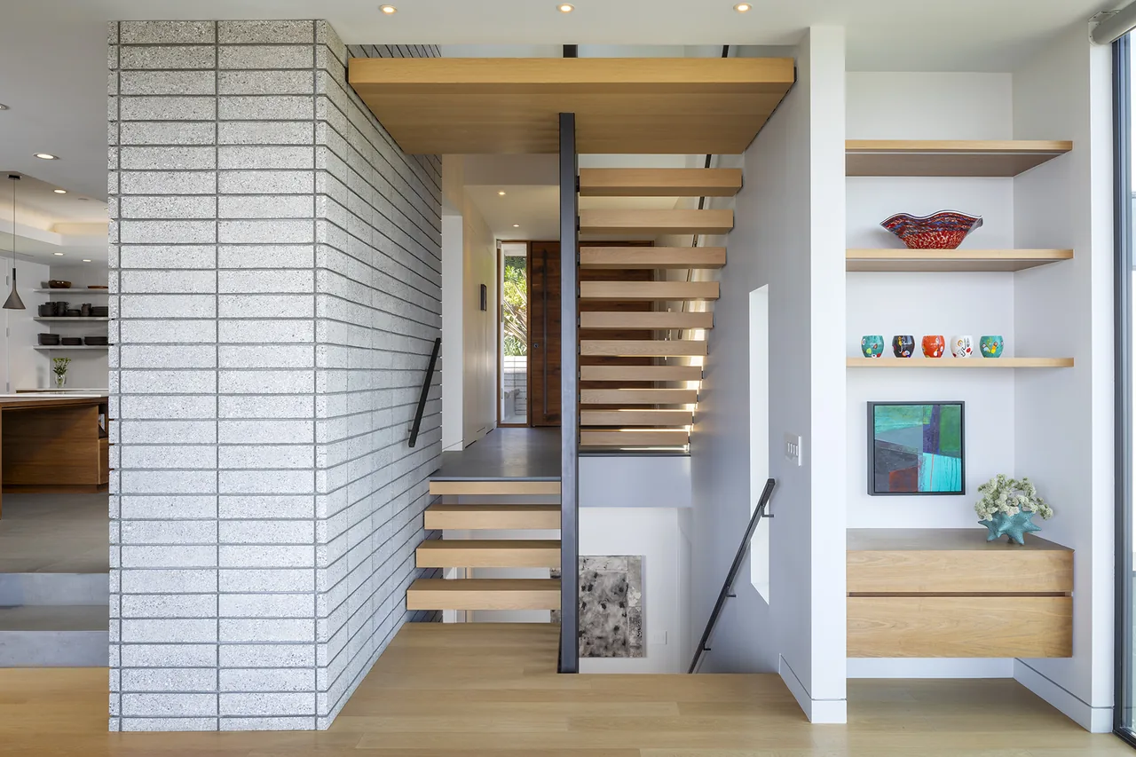 Modern staircase with wood treads and black metal balusters adjacent to a textured gray brick wall and built-in shelves.