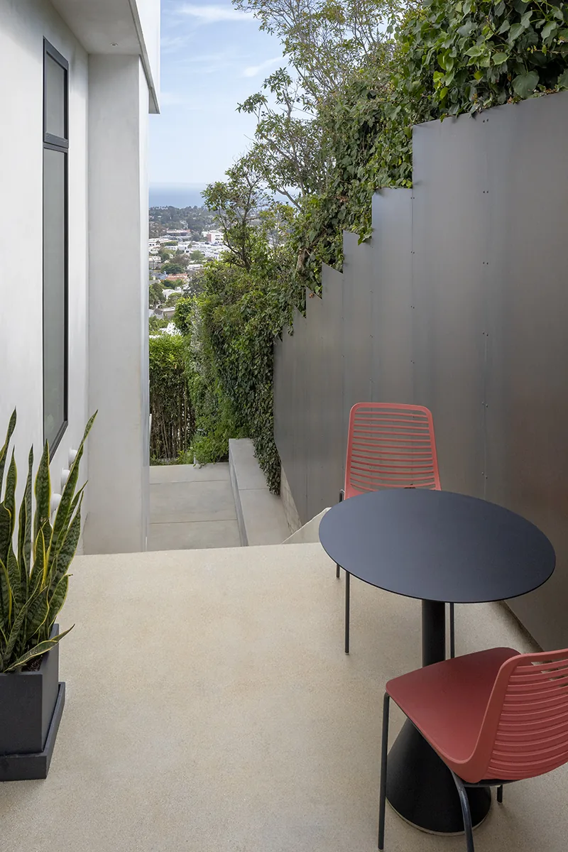 Outdoor deck with red metal chairs and black round table overlooking landscaped slope and city view at Palisades House by Rob Paulus Architects.