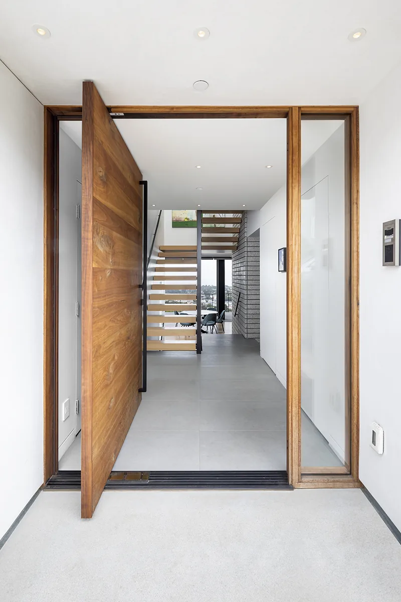 Entry view through a large wooden pivot door showing a floating staircase and dining chairs beyond.