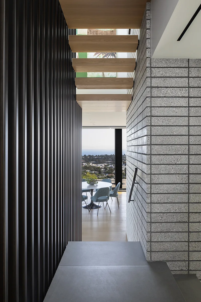Interior stairwell with wood floating treads and black vertical wall slats leads to a dining area with views outside.