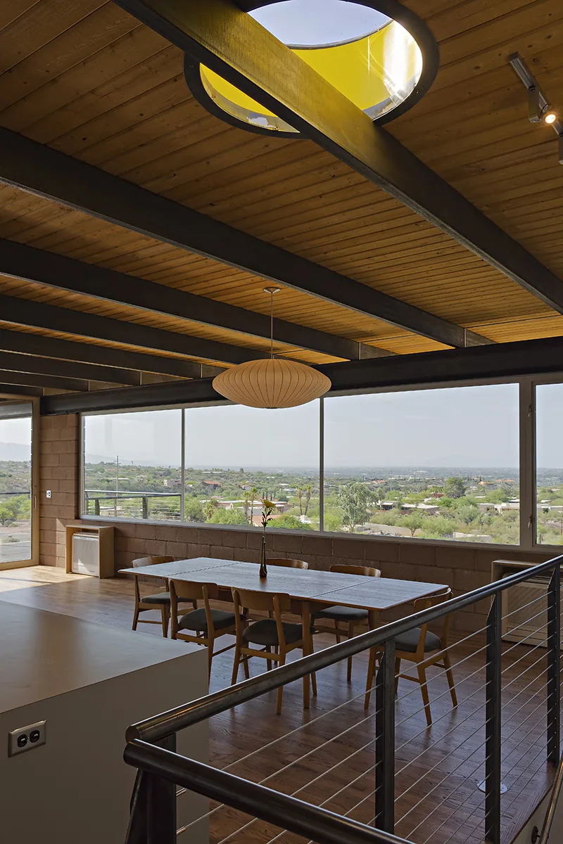 Dining space inside DWC House with wood ceiling beams, cable railing, and expansive desert views seen through large windows.