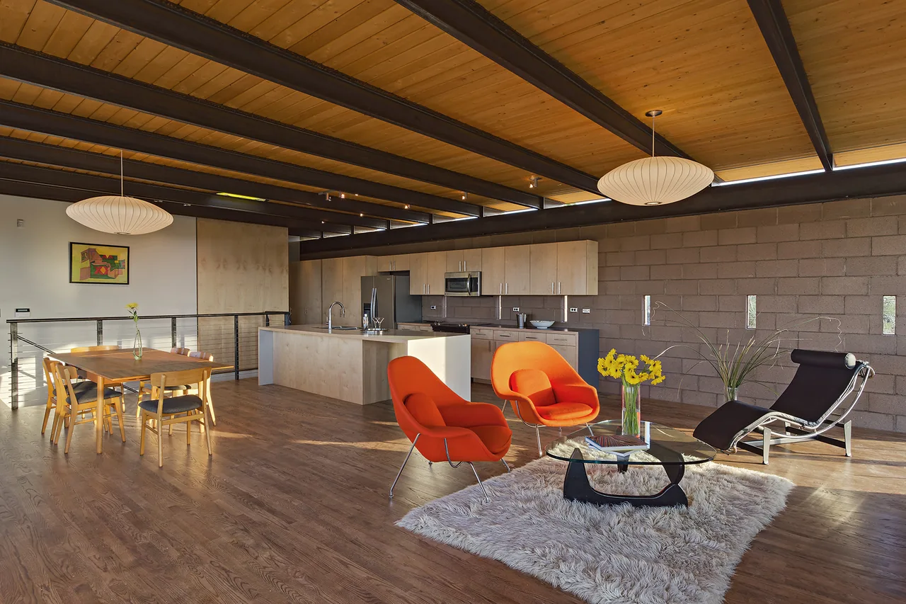 Open-plan living and kitchen in DWC House shows wood ceiling planks and exposed beams with modern furnishings and concrete block walls.