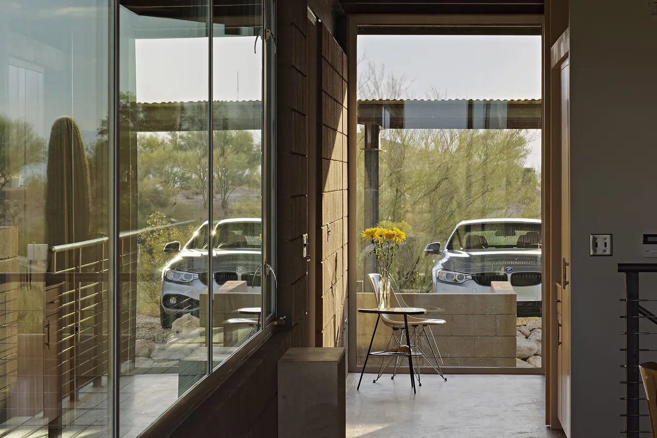 Glass door and window in a desert home opens to a view of desert vegetation and a parked car outside the patio.