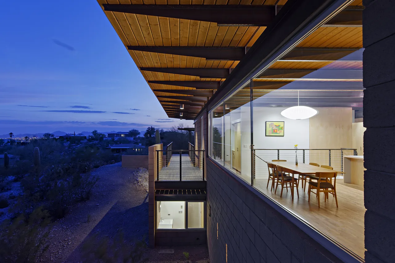 DWC House by Rob Paulus Architects features a wood-paneled ceiling and floor-to-ceiling windows overlooking desert landscape at dusk.