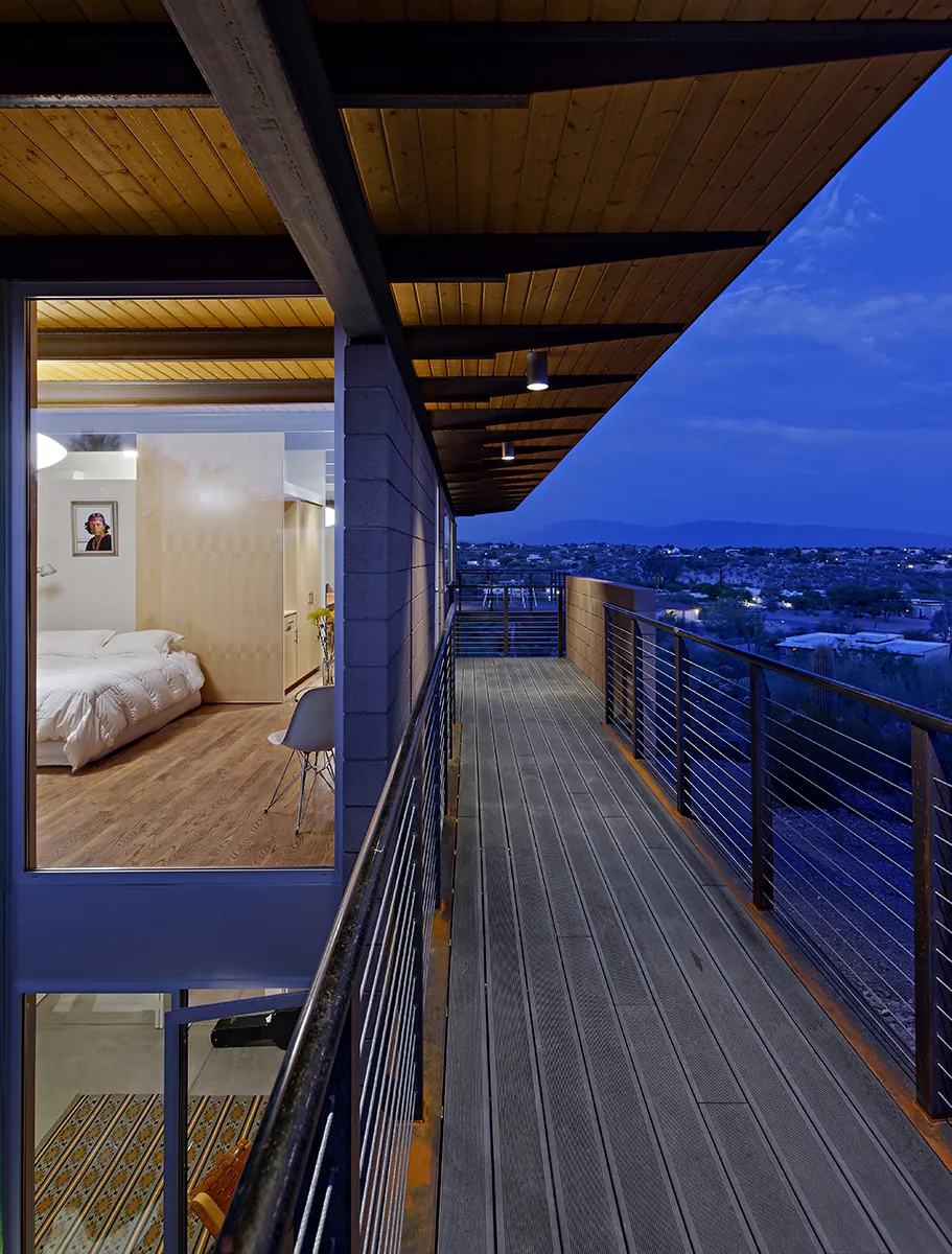 Balcony at DWC House by Rob Paulus Architects with cable railing, wood ceiling, and cinder block wall at dusk.