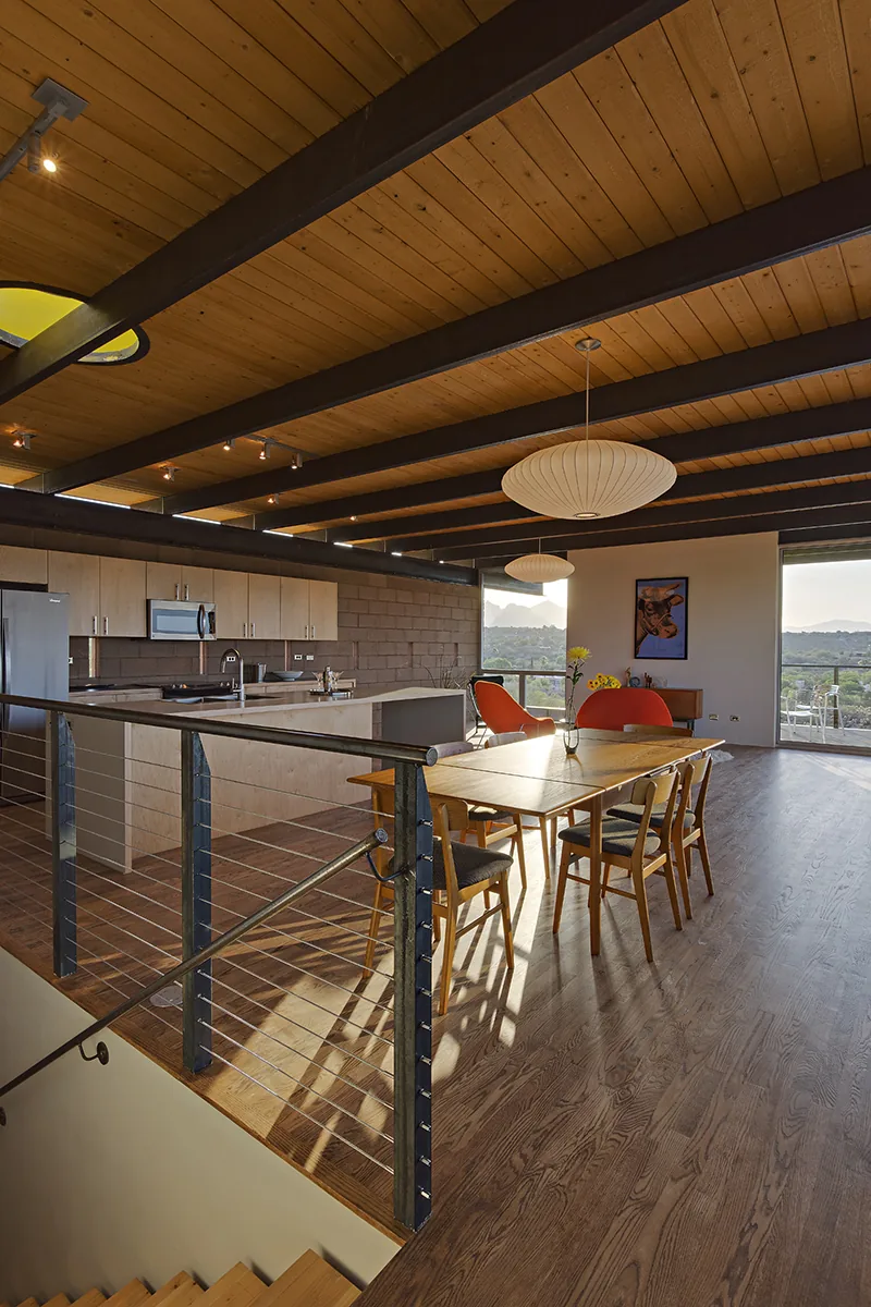 DWC House dining space features a wooden table, exposed beam ceiling, and steel railing overlooking the kitchen area and scenic views.