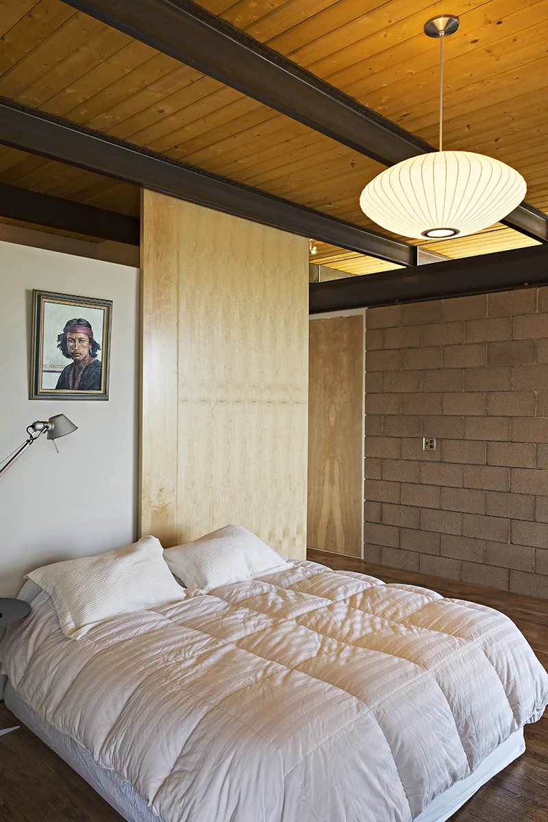 A bedroom in the DWC House features exposed wood beams, a wood ceiling, and a cinder block wall.