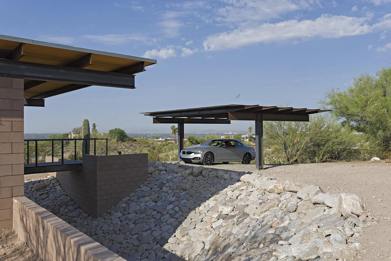 Carport with corrugated metal roof and steel supports near a rocky slope and bridging structure at DWC House.