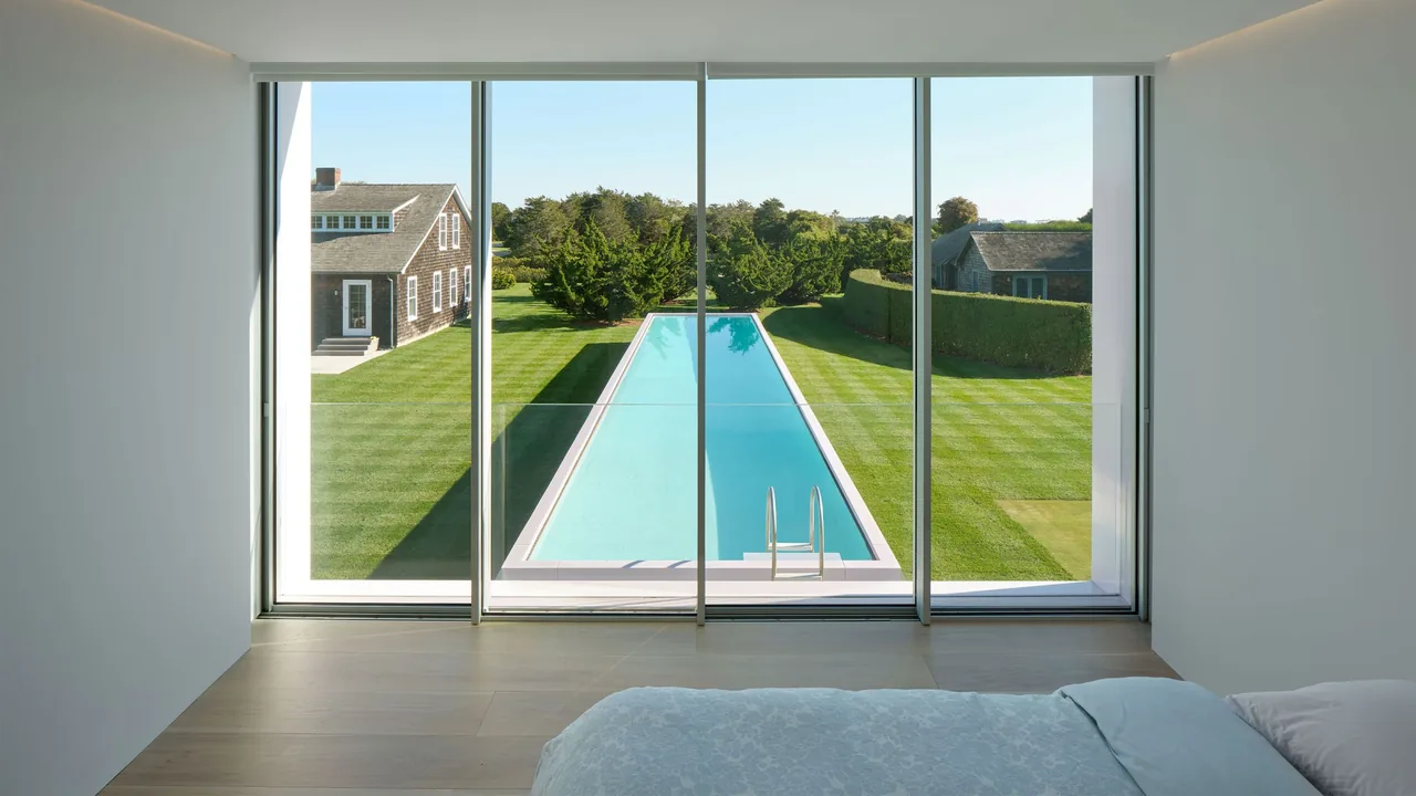 Minimalist bedroom with floor-to-ceiling glass walls overlooking a rectangular pool and manicured lawn.