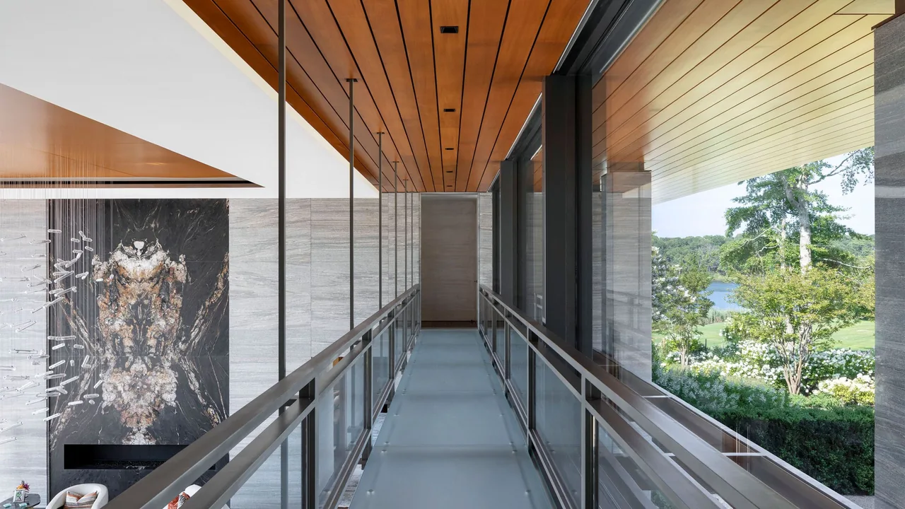 Modern interior corridor with glass railing and wood ceiling leading to a light-filled space overlooking greenery and water.