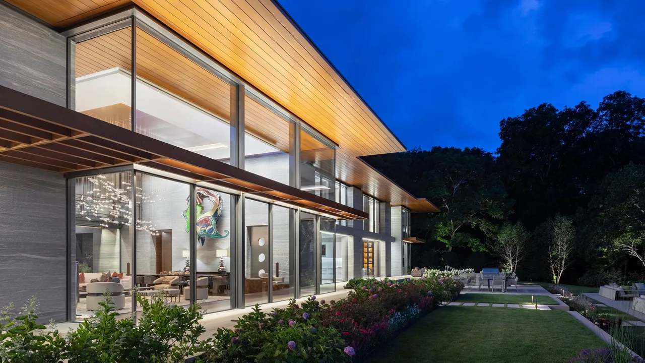 Modern home with extensive glazing under an overhanging wood-paneled roof overlooking a landscaped outdoor seating area and garden.