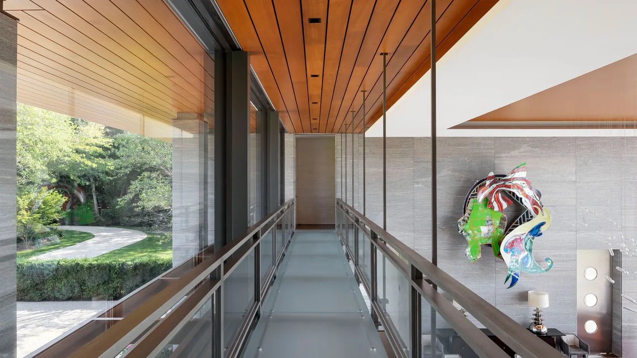 Glass hallway with wood-paneled ceiling above a living area featuring a colorful abstract wall sculpture at Millstone project.