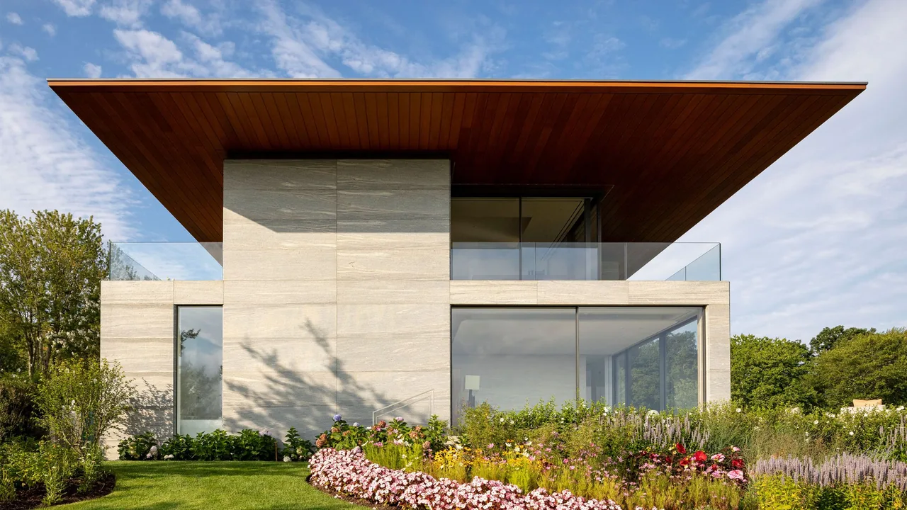 Millstone house features a flat, overhanging wood ceiling roof above smooth stone walls and large glass windows overlooking a garden.