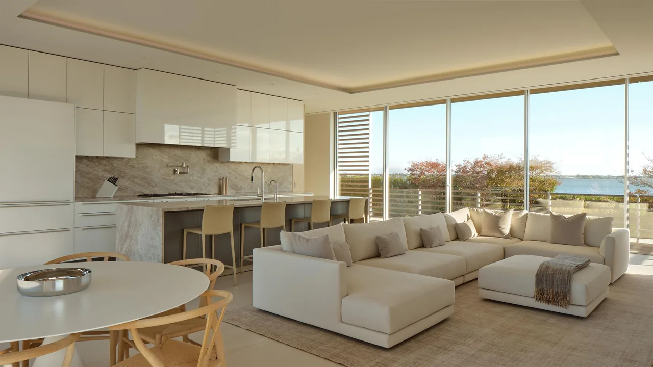 Open living room with sectional sofa and kitchen area featuring a large island and seating, overlooking a scenic waterside view.