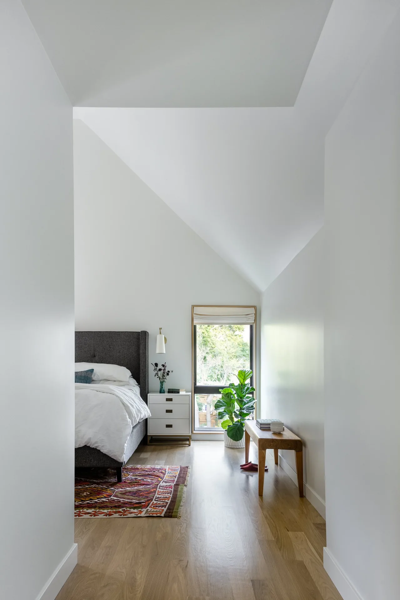 A minimalist bedroom with an angled ceiling, gray upholstered bed, wooden floors, and a decorative rug near a tall window.