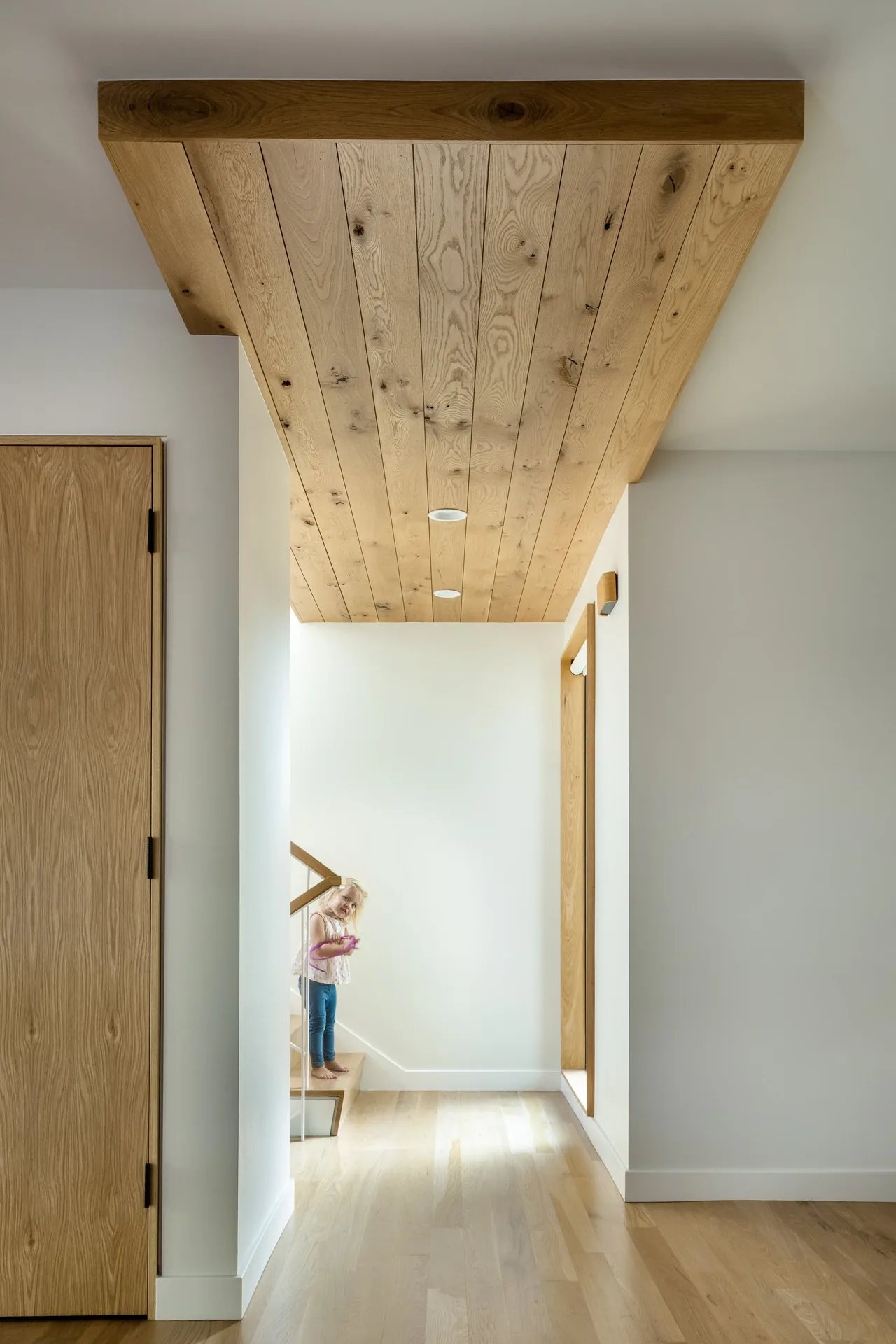 Hallway with wood plank ceiling and light oak flooring featuring wooden doors and stairs with a child standing.