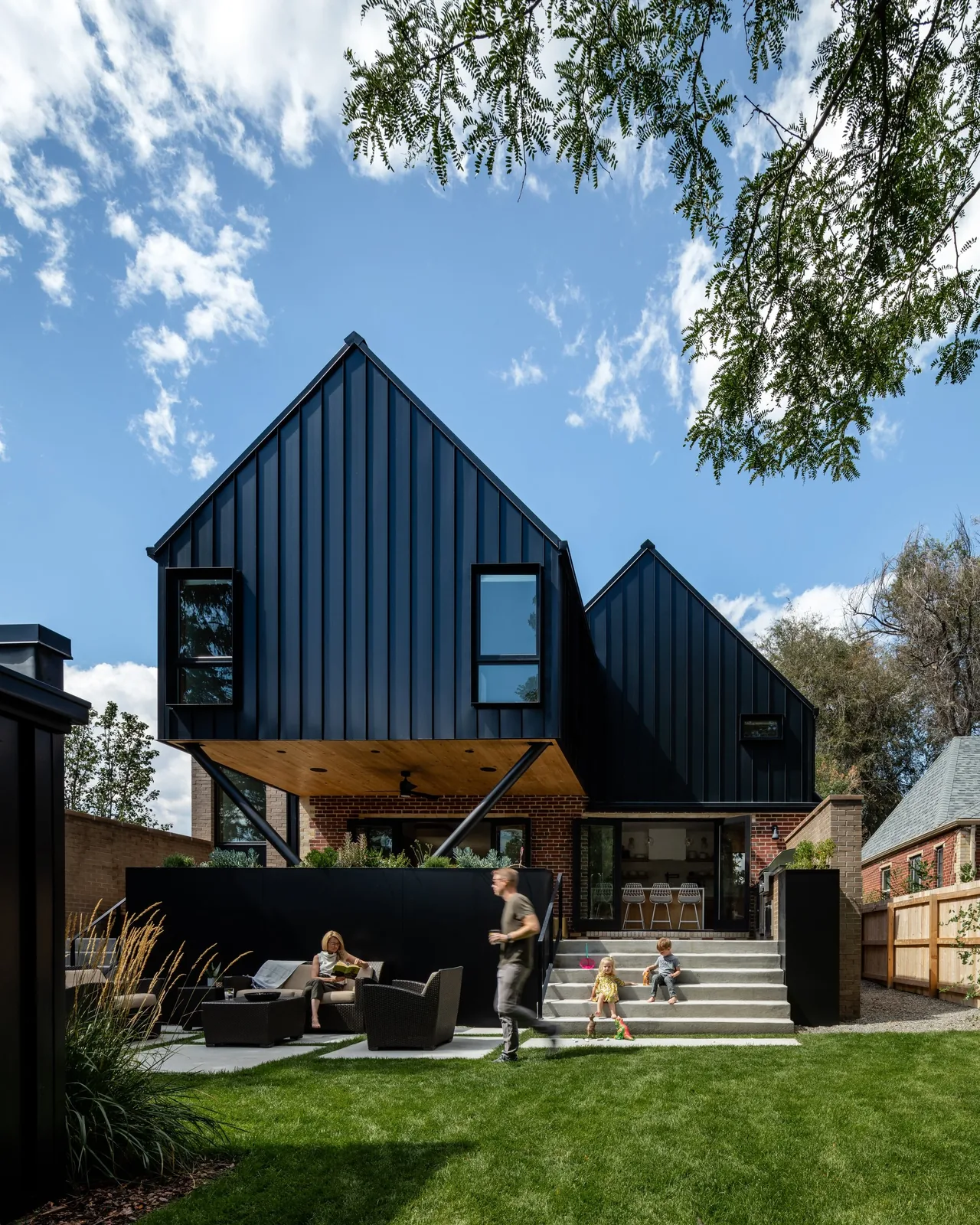 Two-story home with black metal cladding above brick in the backyard, featuring outdoor seating and a grass lawn.