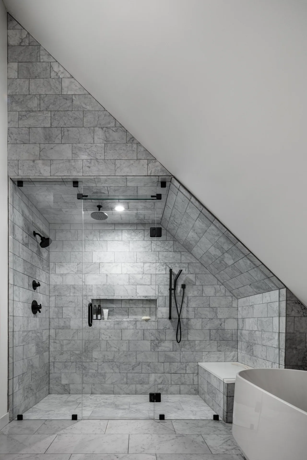 Shower area in a modern bathroom with marble tile walls, black shower fixtures, and a glass enclosure under a sloped ceiling.