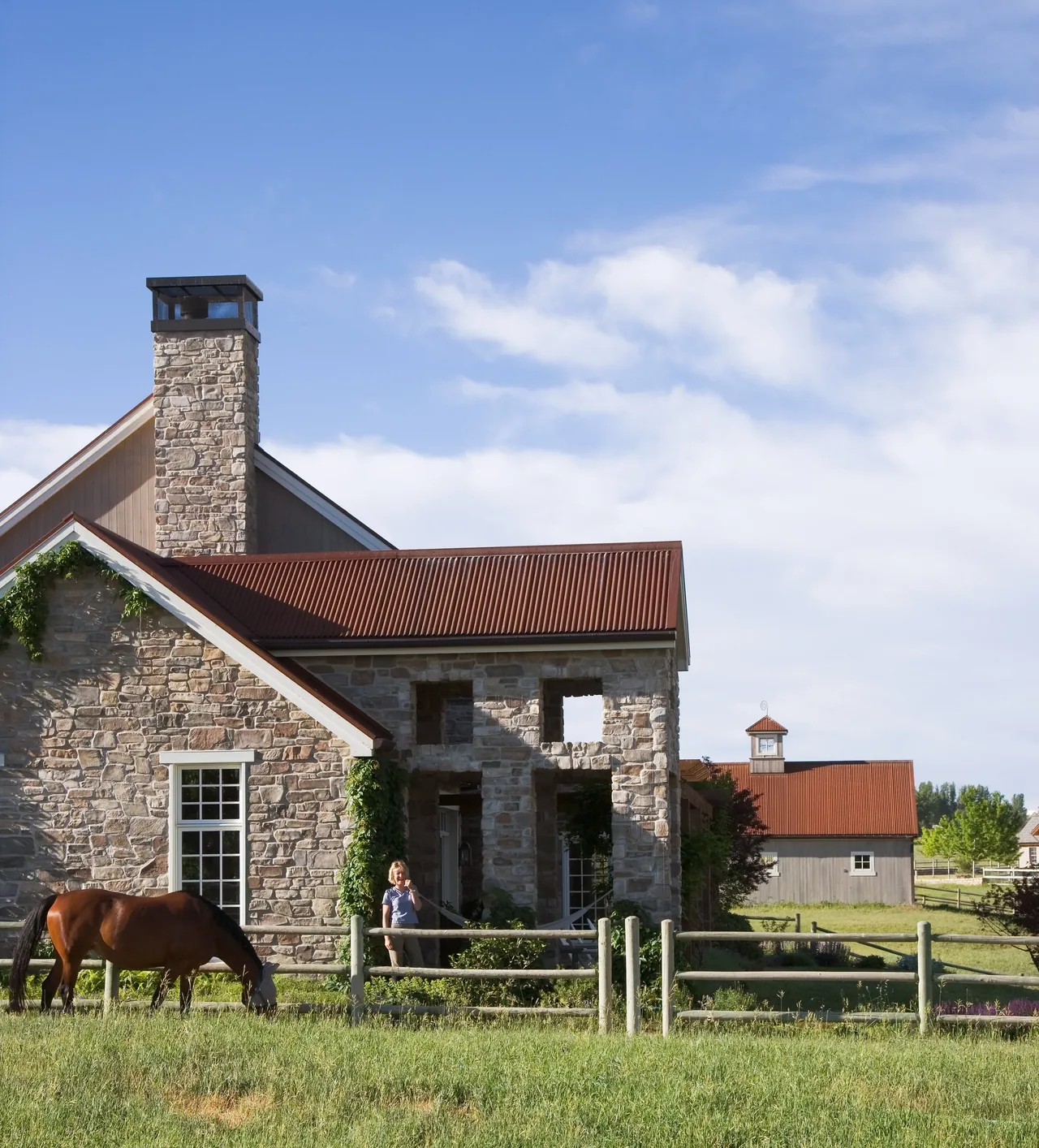Creekside Compound features a stone house with red metal roof and a barn with a cupola in the background.