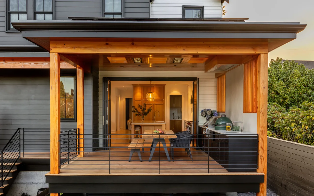 Covered patio with wooden framing, black railing, outdoor kitchen, and dining table opening to interior kitchen.