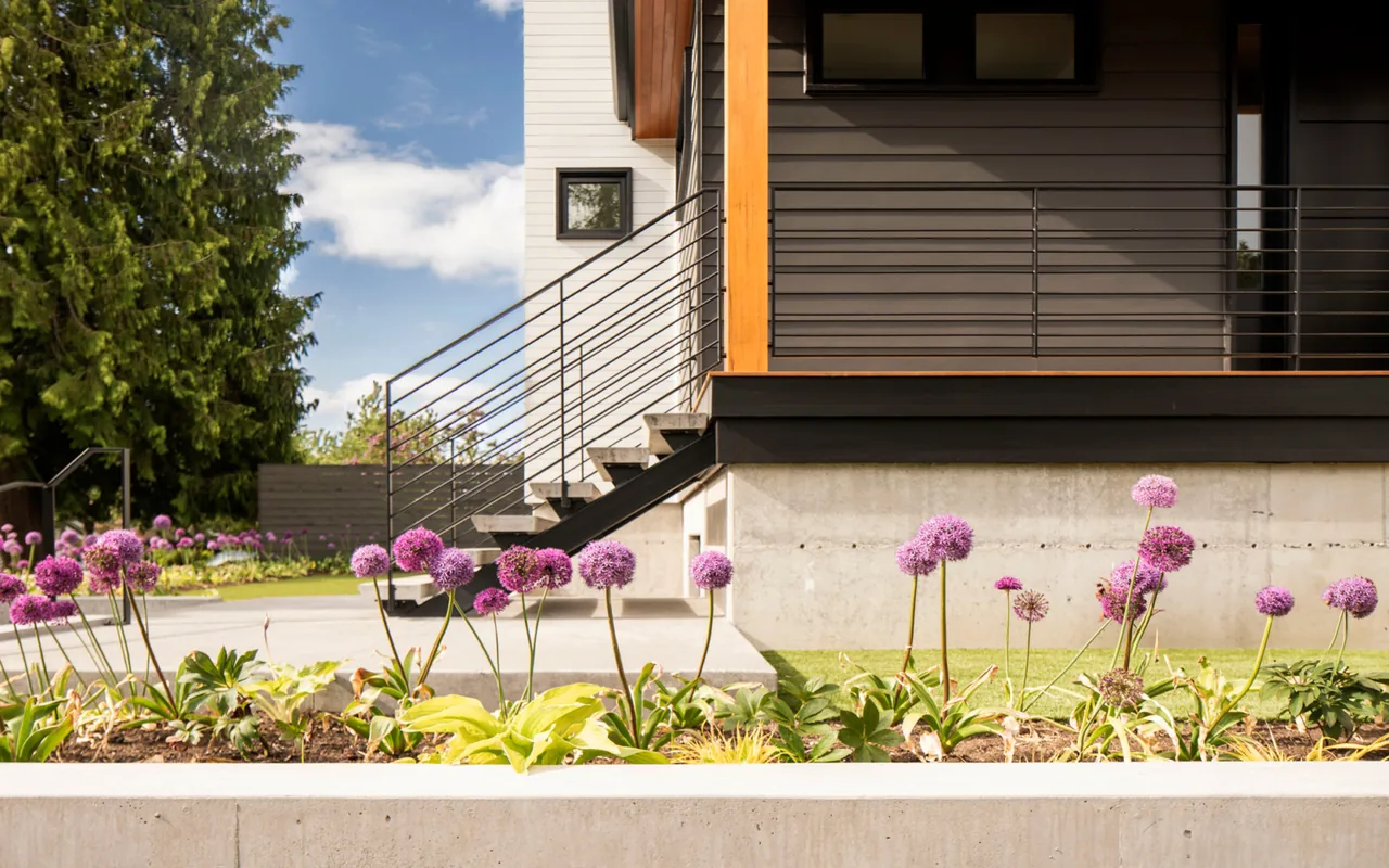 Exterior detail of a house featuring black metal stair railing, mixed siding colors, and a flower bed with purple blooms.