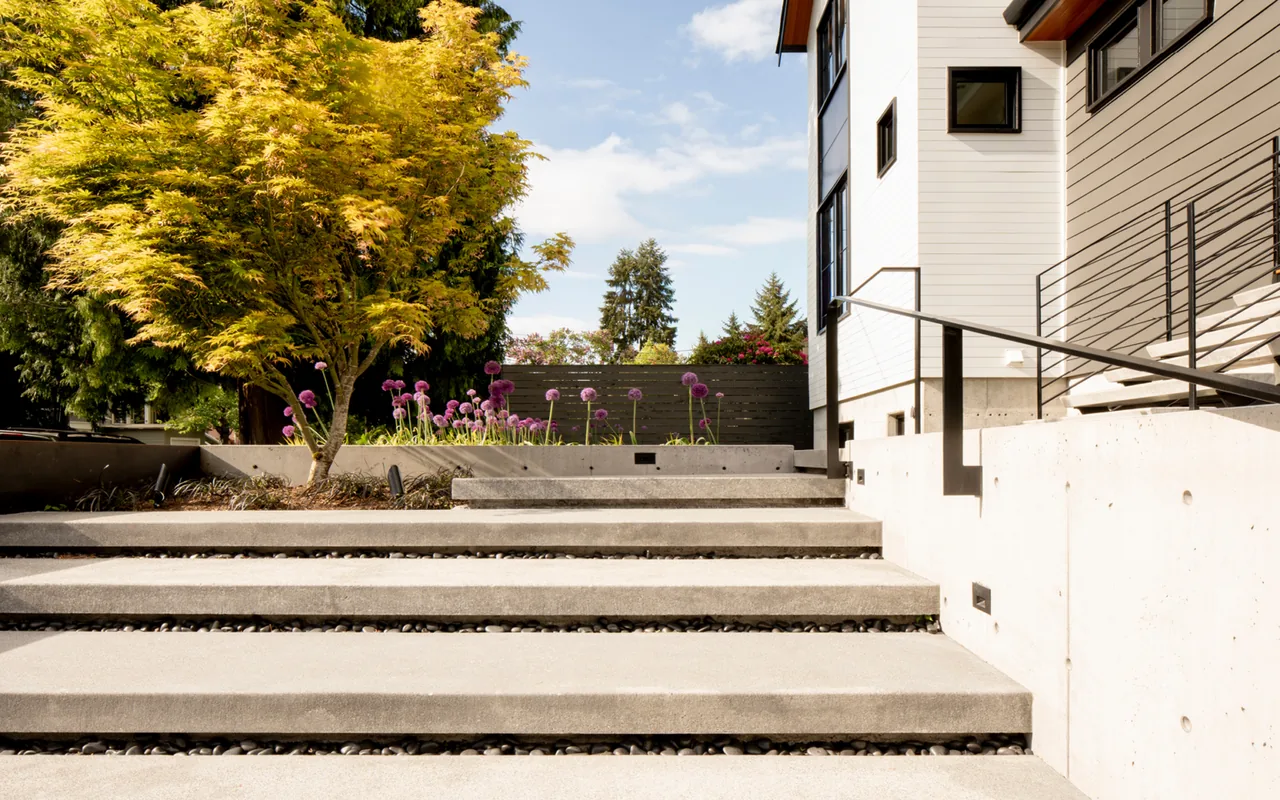 Concrete stairs with black metal railing lead up to a modern residential entry surrounded by landscaping and trees.