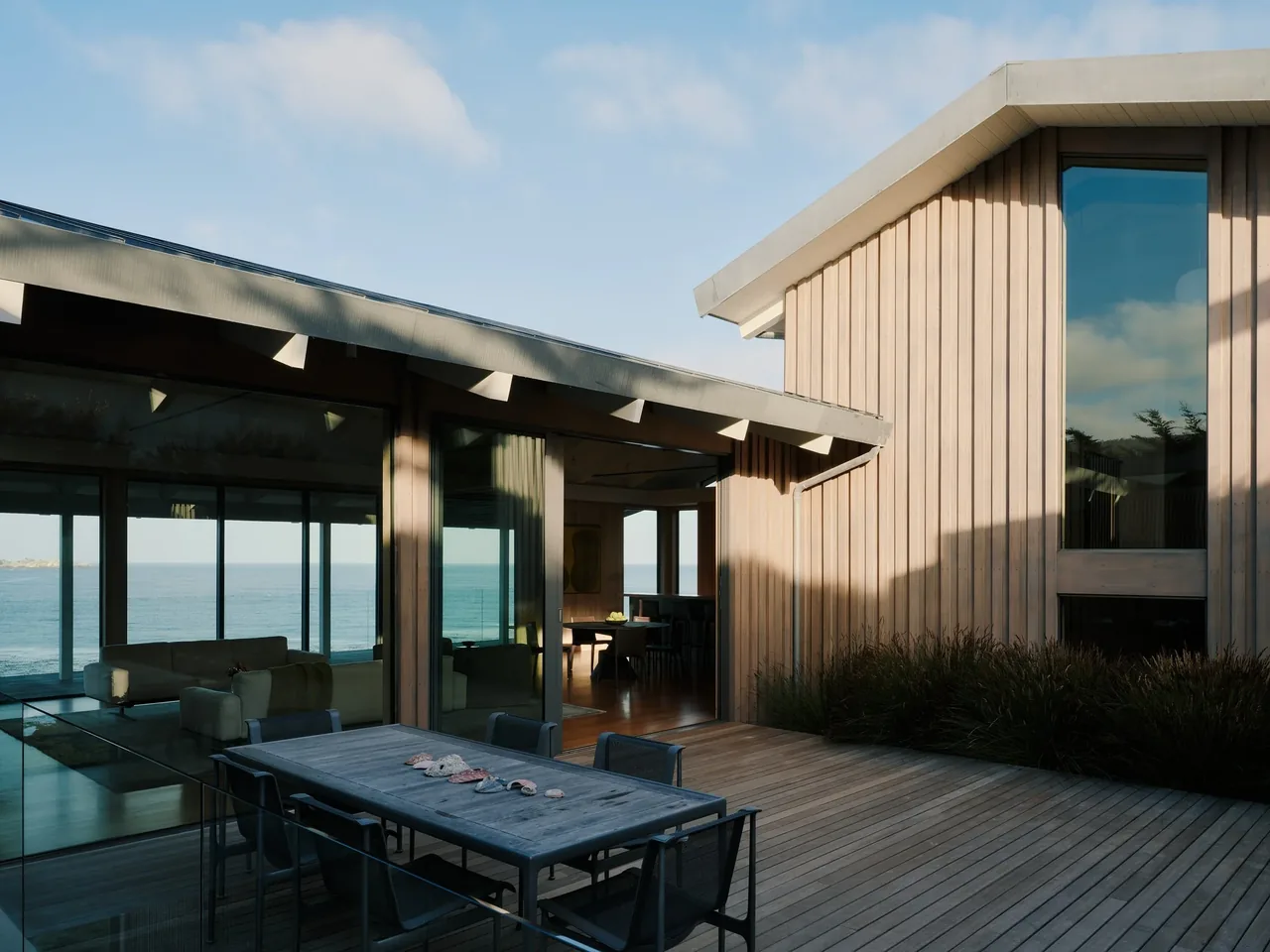 The Carmel project features vertical wood siding and a wood deck with outdoor dining, overlooking coastal waters.